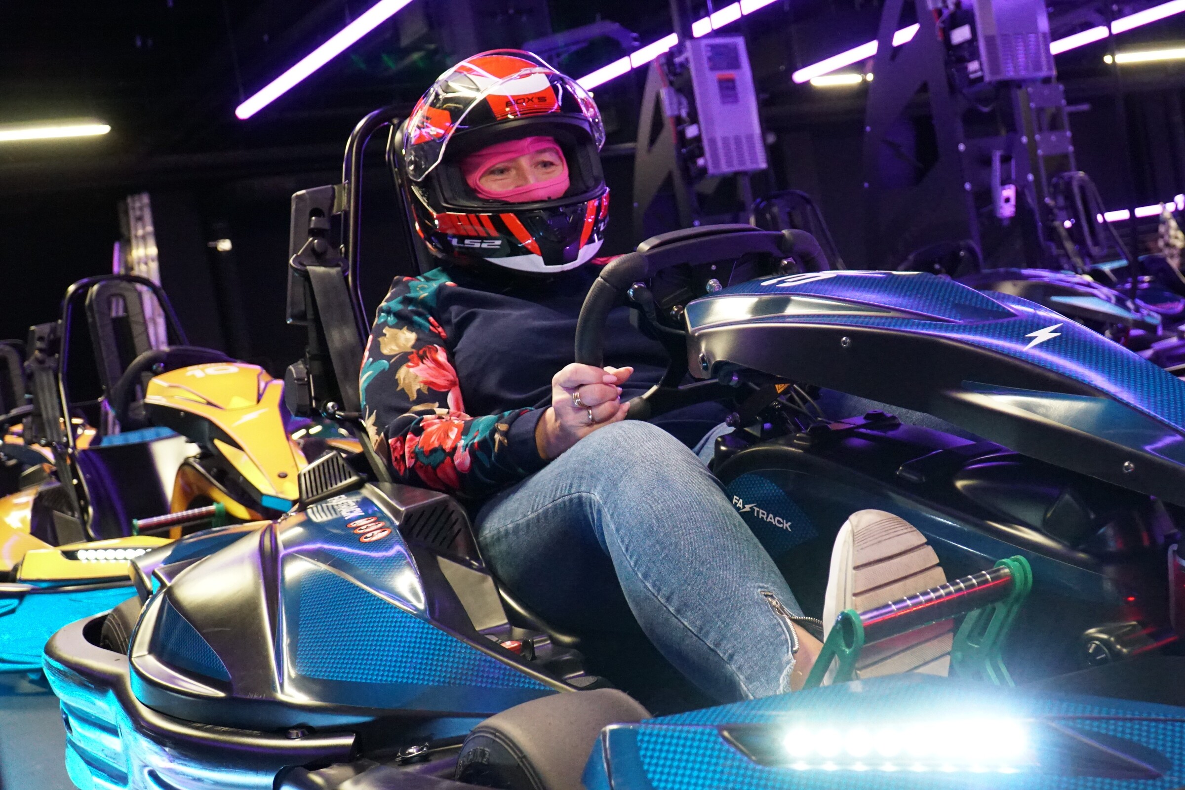 Kobieta w kasku siedzi w gokarcie na torze Fast Track, trzymając kierownicę i uśmiechając się. W tle inne pojazdy i oświetlenie toru.